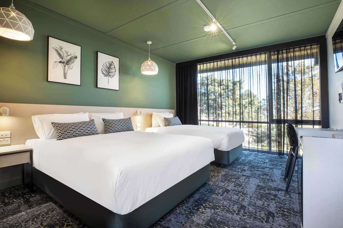 A hotel room with two beds, a large window with curtains, and artwork on the wall.