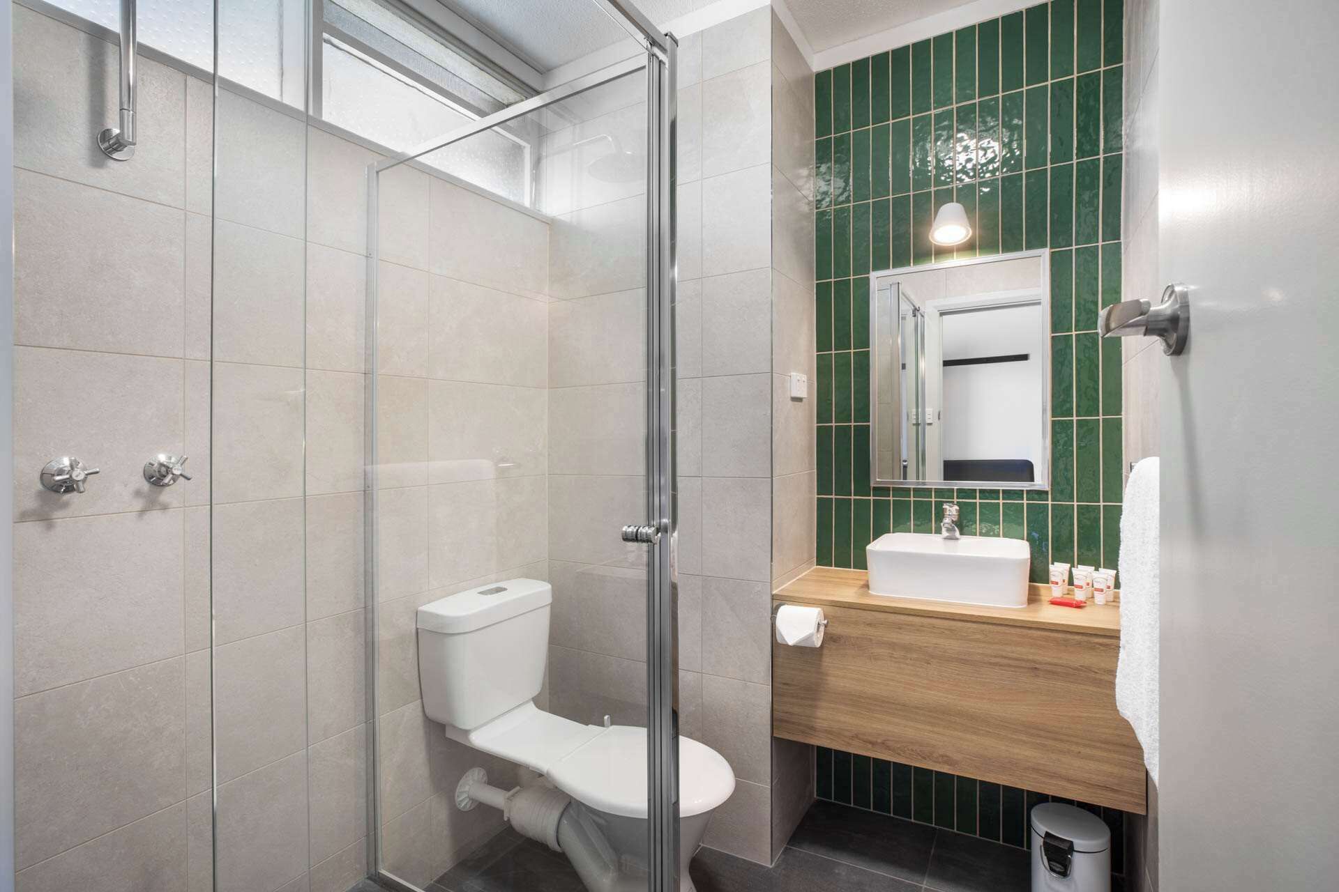 Un baño moderno con cabina de ducha de vidrio, inodoro, lavabo con espejo y pared de azulejos verdes.