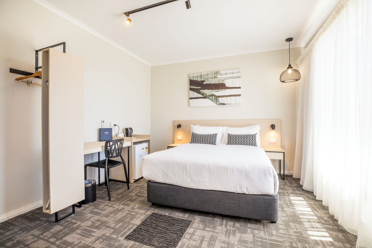 A hotel room with a bed, desk, chair, and a painting on the wall.