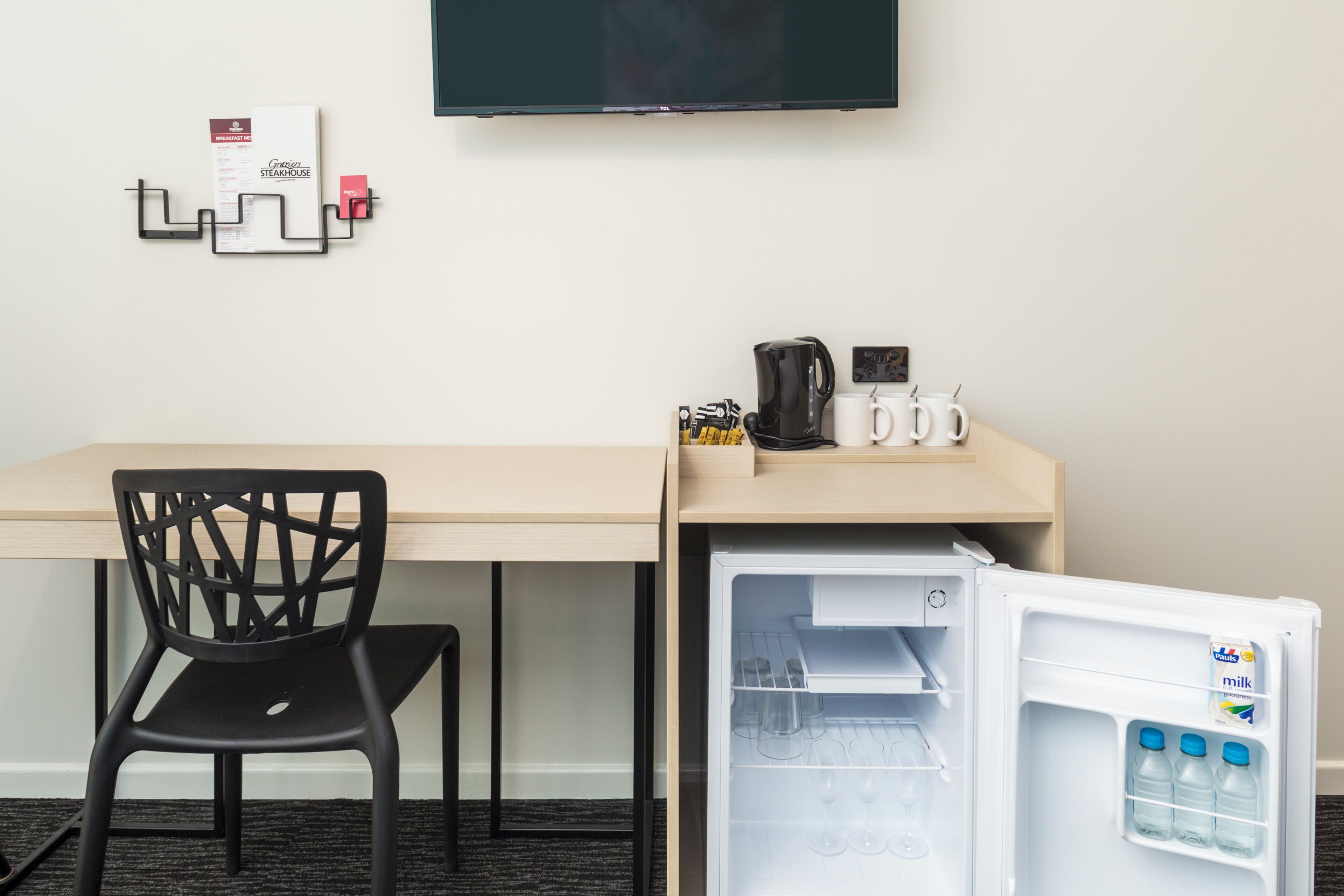 A hotel room with a desk, a chair, a flat-screen TV mounted on the wall, and a mini-fridge with milk bottles.