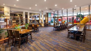 A spacious indoor area with multiple dining tables and chairs, a staircase, and large windows offering views of outdoor play equipment.