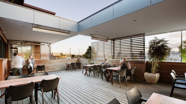 An outdoor patio area with wooden flooring, tables, and chairs. There are large windows with blinds, and a view of a street with cars and buildings.