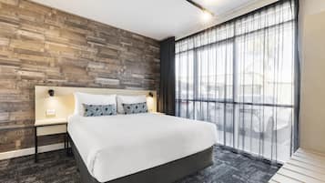 A hotel room with a large bed, a wooden wall with numbers, a nightstand, and a window with sheer curtains.