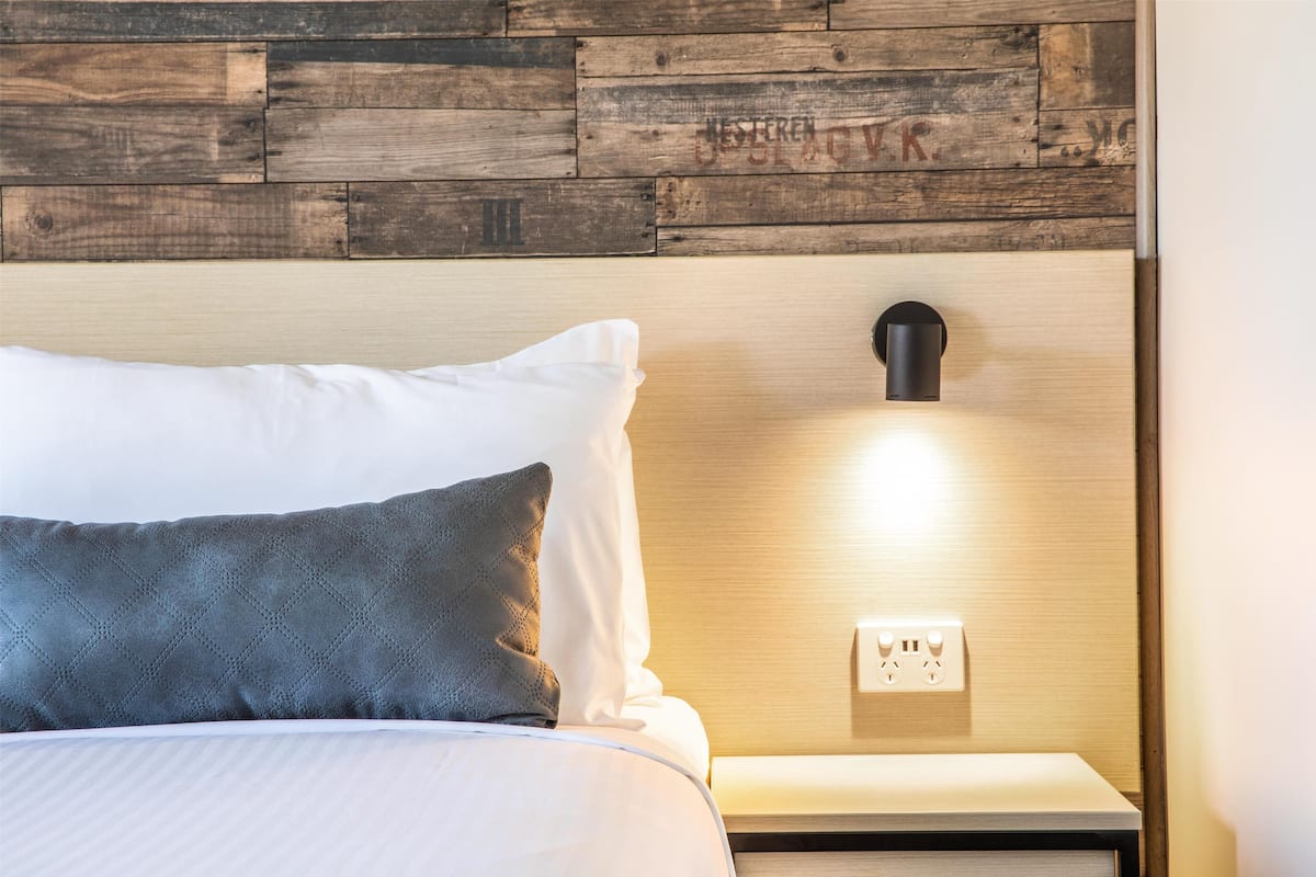 A neatly made bed with white and dark grey pillows, a wooden headboard, and a bedside lamp.