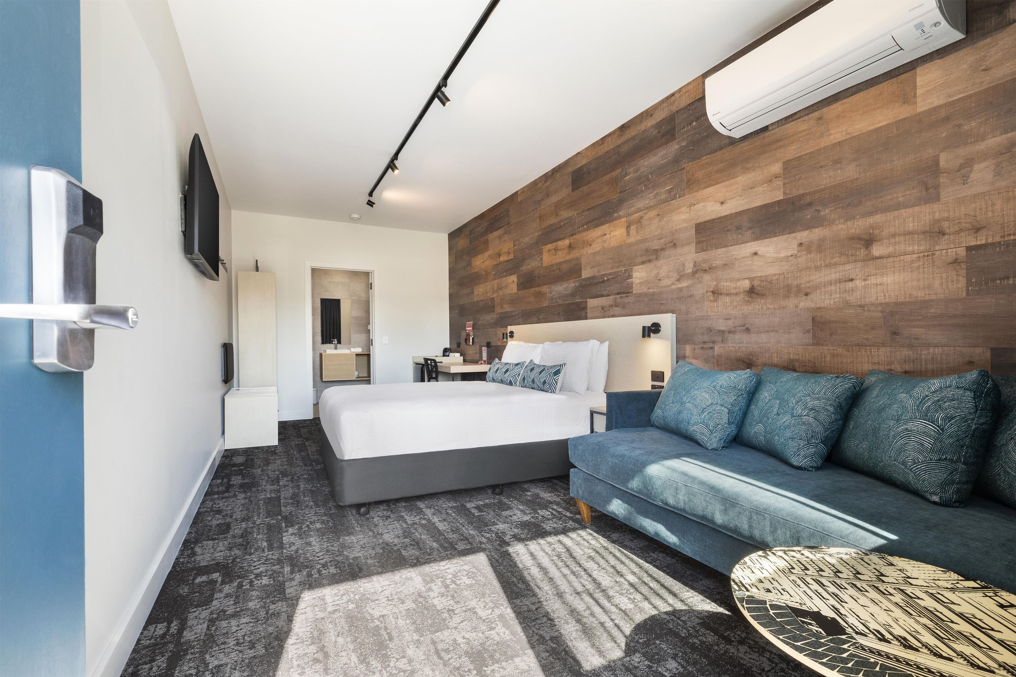 A modern hotel room with a bed, a sofa, a TV, and an air conditioning unit.