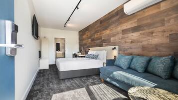 A modern hotel room with a bed, a sofa, a TV, and an air conditioning unit.