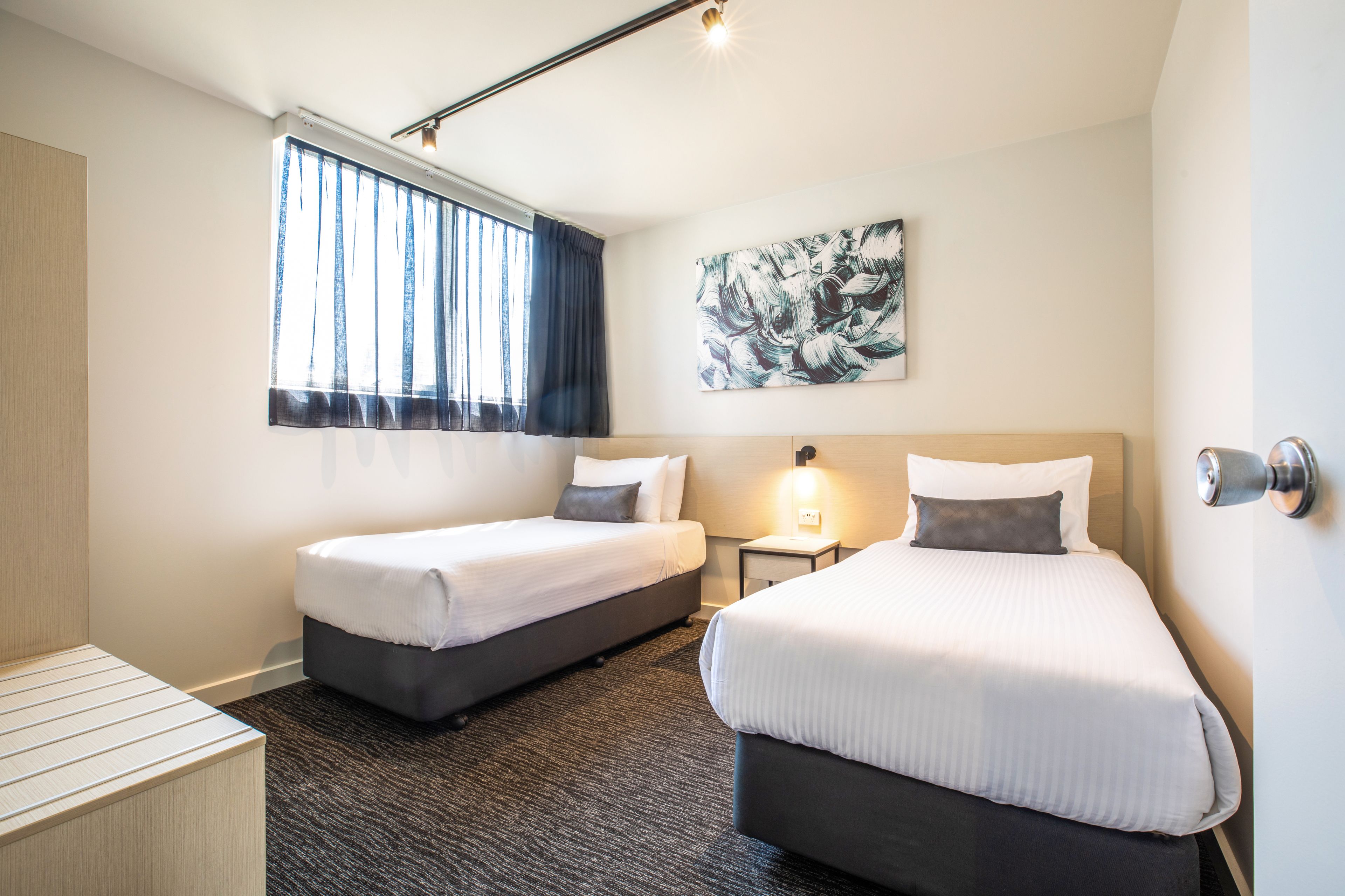Ein Hotelzimmer mit zwei Betten, einem Schreibtisch, einem Nachttisch, einem Fenster mit Vorhängen und einem Wandbild.