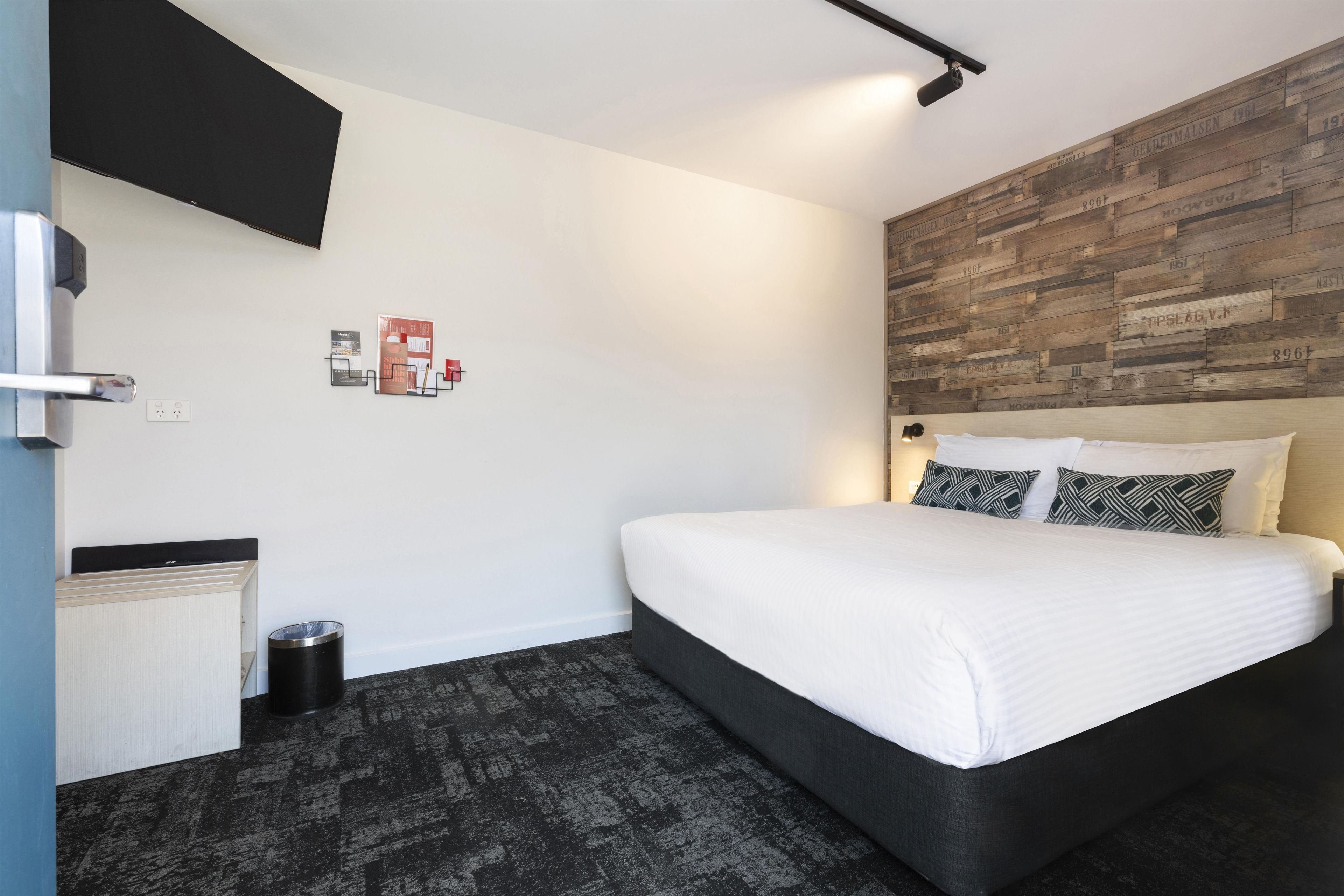 A modern hotel room with a bed, a flat-screen TV, a wooden accent wall, and a small trash bin.