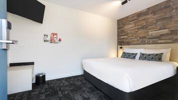 A modern hotel room with a bed, a flat-screen TV, a wooden accent wall, and a small trash bin.