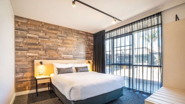 A modern hotel room with a large bed, a wooden accent wall, a nightstand with a lamp, and a view of the outdoors through large windows. - Nightcap at Westside Hotel (Laverton North)