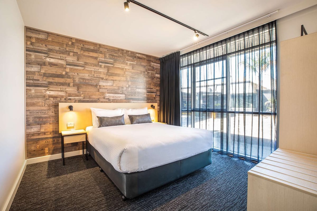 A modern hotel room with a large bed, a wooden accent wall, a nightstand with a lamp, and a view of the outdoors through large windows.