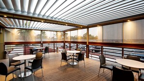 An outdoor patio area with round tables and chairs, a wooden deck, and a glass ceiling with horizontal beams. - Nightcap at Westside Hotel (Laverton North)