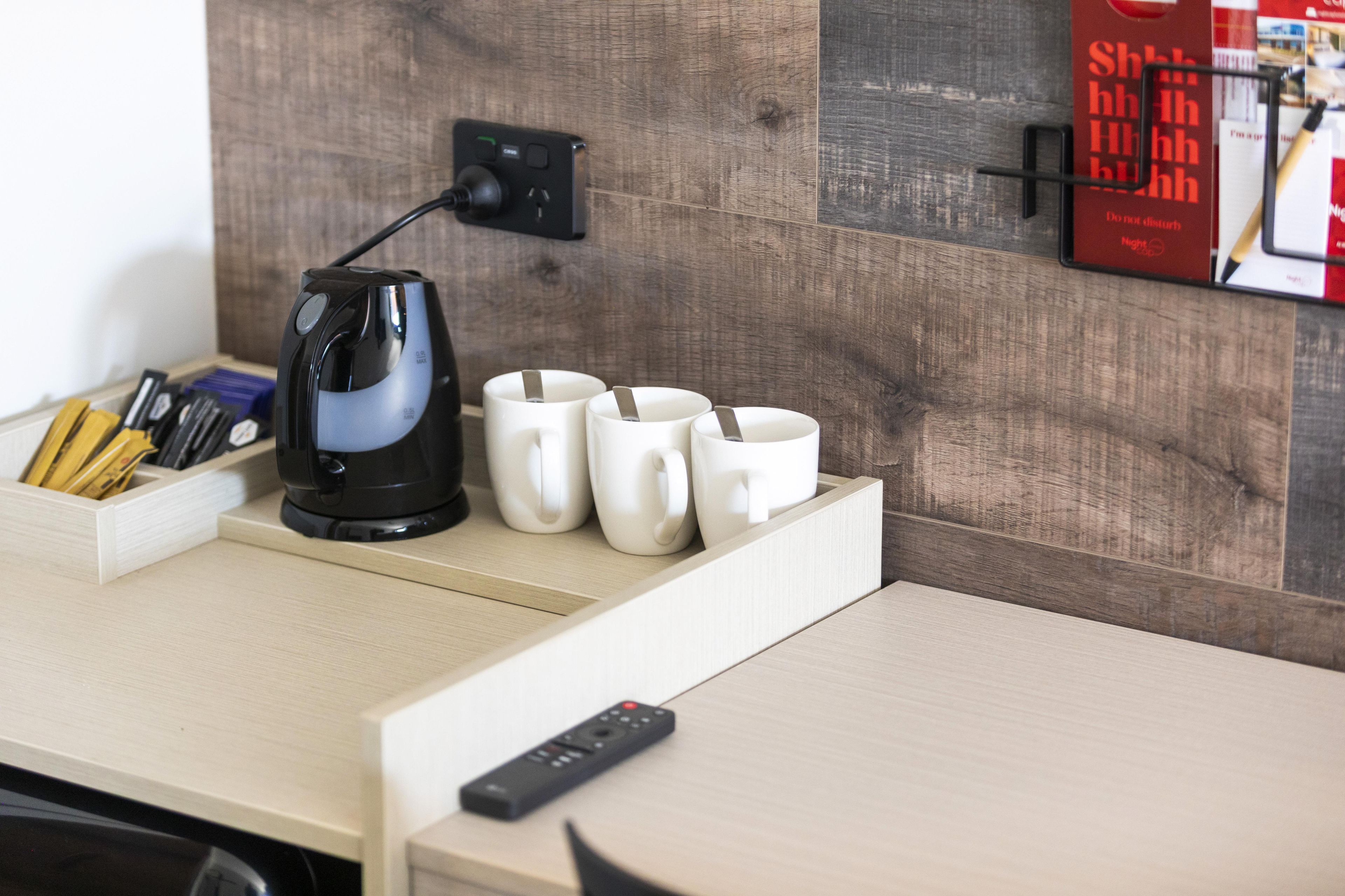 Un escritorio de madera con una cafetera negra, cuatro tazas blancas, un control remoto y un portalápices con bolígrafos.