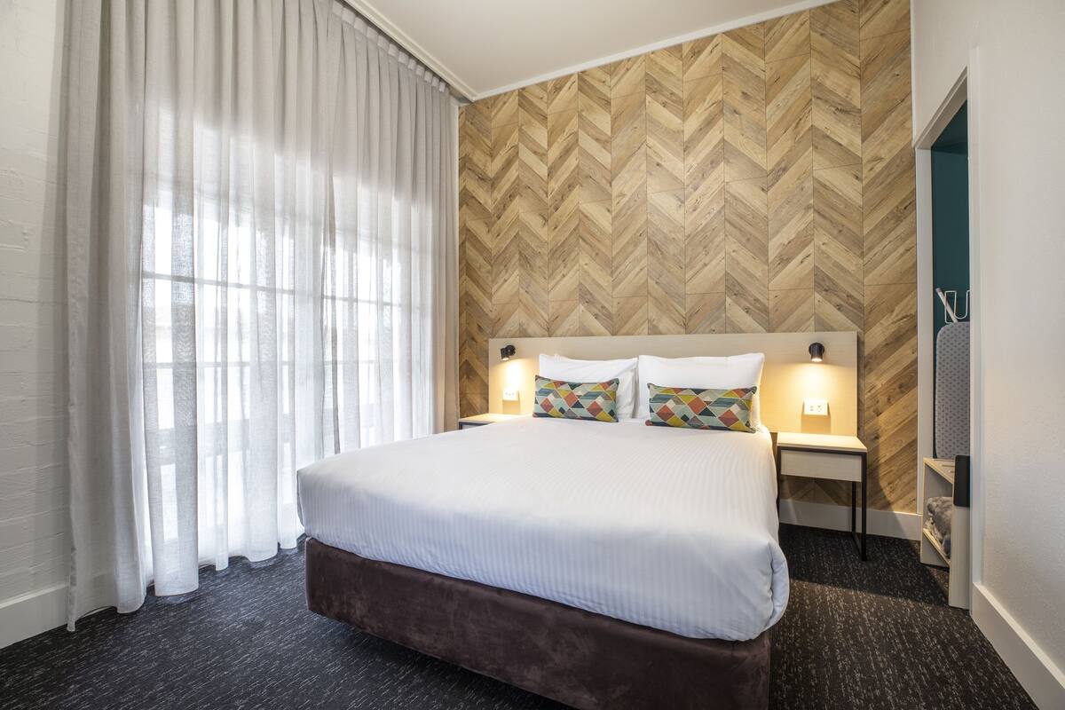 A hotel room with a large bed, a wooden herringbone-patterned wall, a nightstand with a lamp, and a window with sheer curtains.