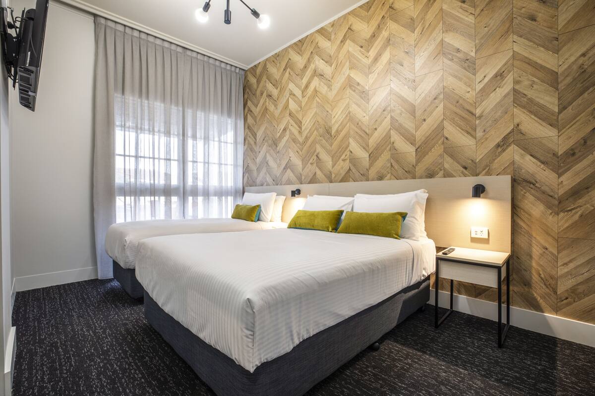 A hotel room with a large bed, a nightstand, a wall-mounted TV, and a window with sheer curtains.