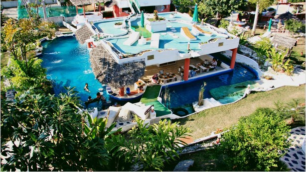 A pool area with multiple sections, including a central pool and a smaller pool connected by a bridge. There are lounge chairs and umbrellas around the pools, and a building with a covered patio area. - Mirabel Resort & Mini Golf (Pattaya)