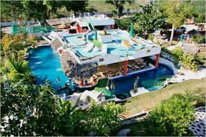 A pool area with multiple sections, including a central pool and a smaller pool connected by a bridge. There are lounge chairs and umbrellas around the pools, and a building with a covered patio area. - Mirabel Resort & Mini Golf (Pattaya)