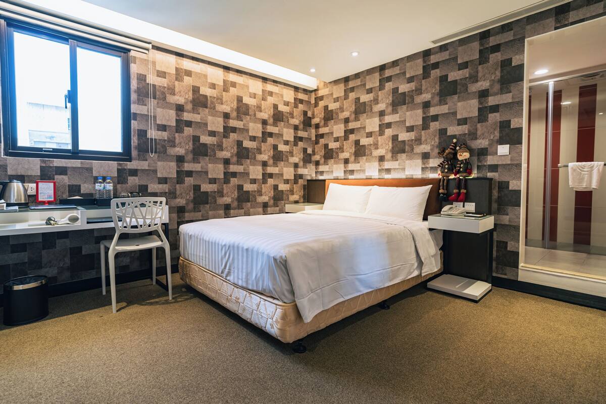 Una habitación de hotel con cama, escritorio, silla, ducha y ventana.