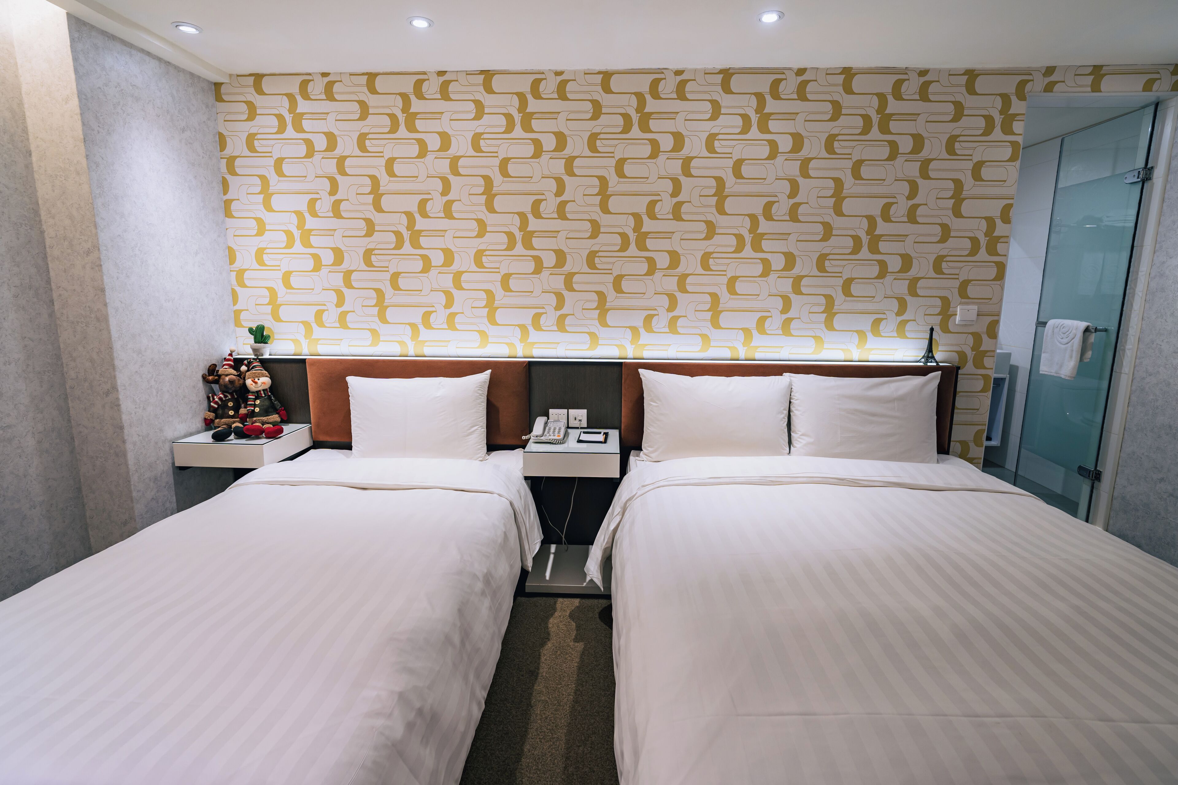 A hotel room with two beds, a nightstand with a phone and a small plant, and a shower area.