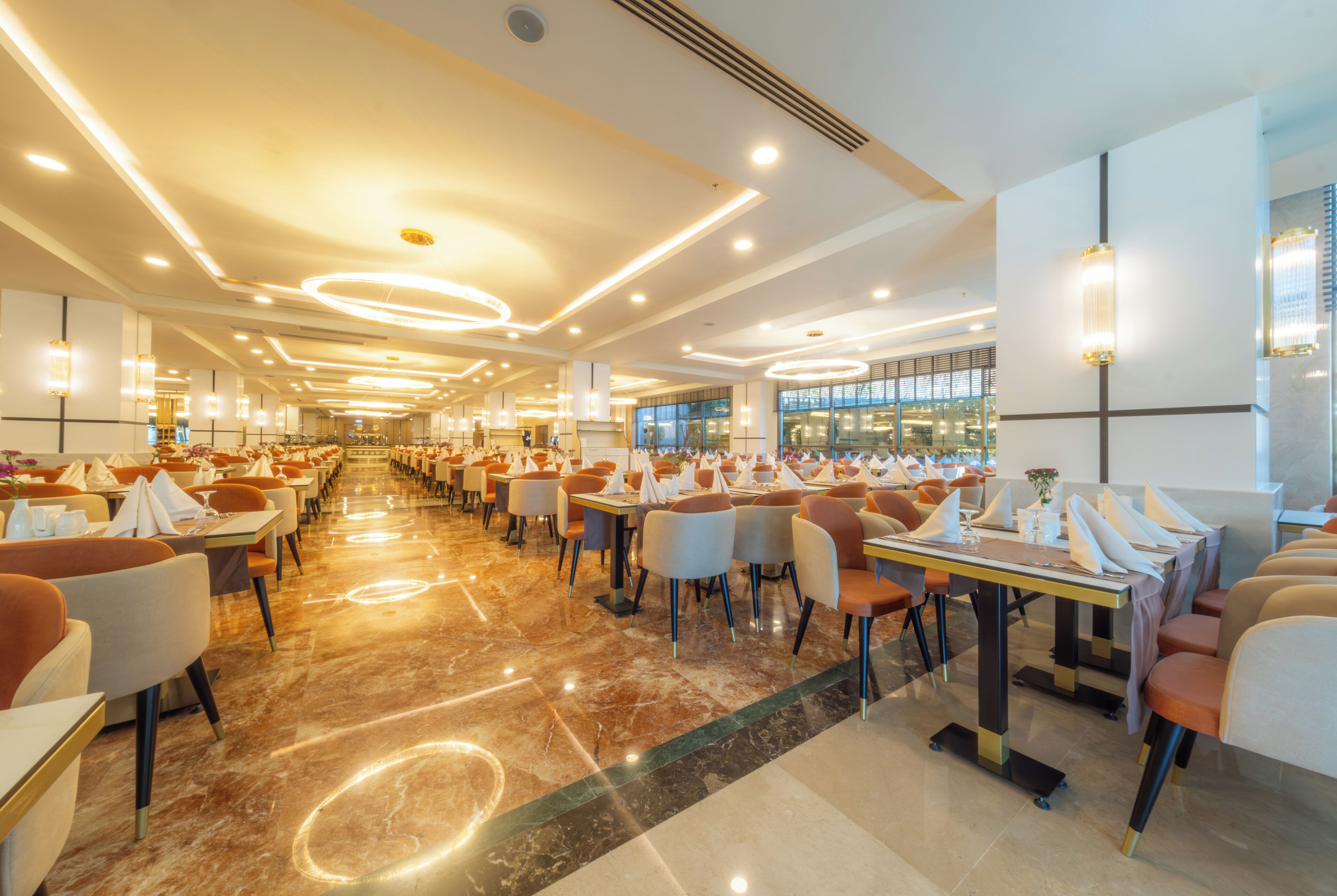 A spacious hotel dining area with multiple tables and chairs, a marble floor, and modern lighting fixtures.