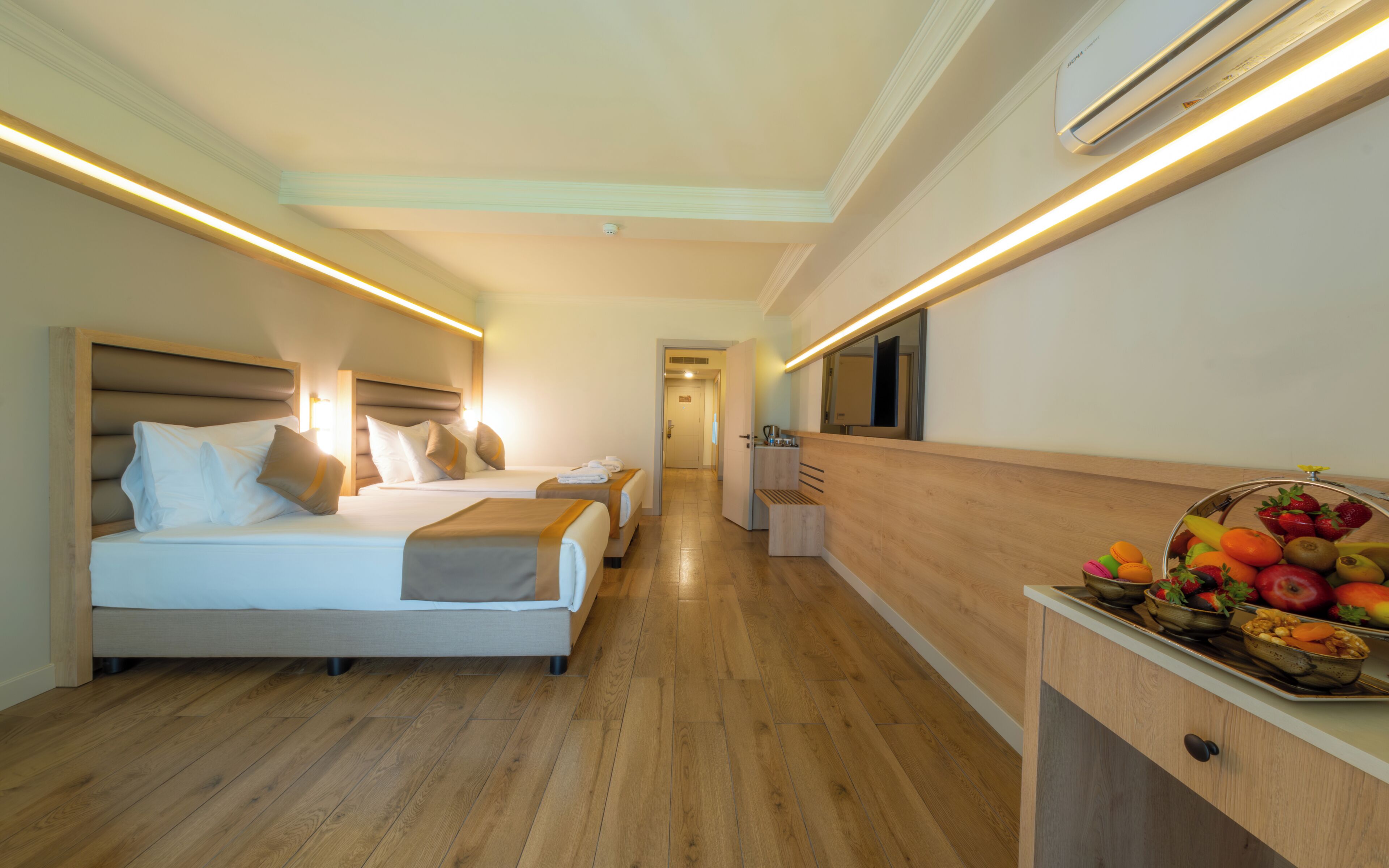 A hotel room with two beds, a wooden floor, a kitchenette, and a fruit basket.