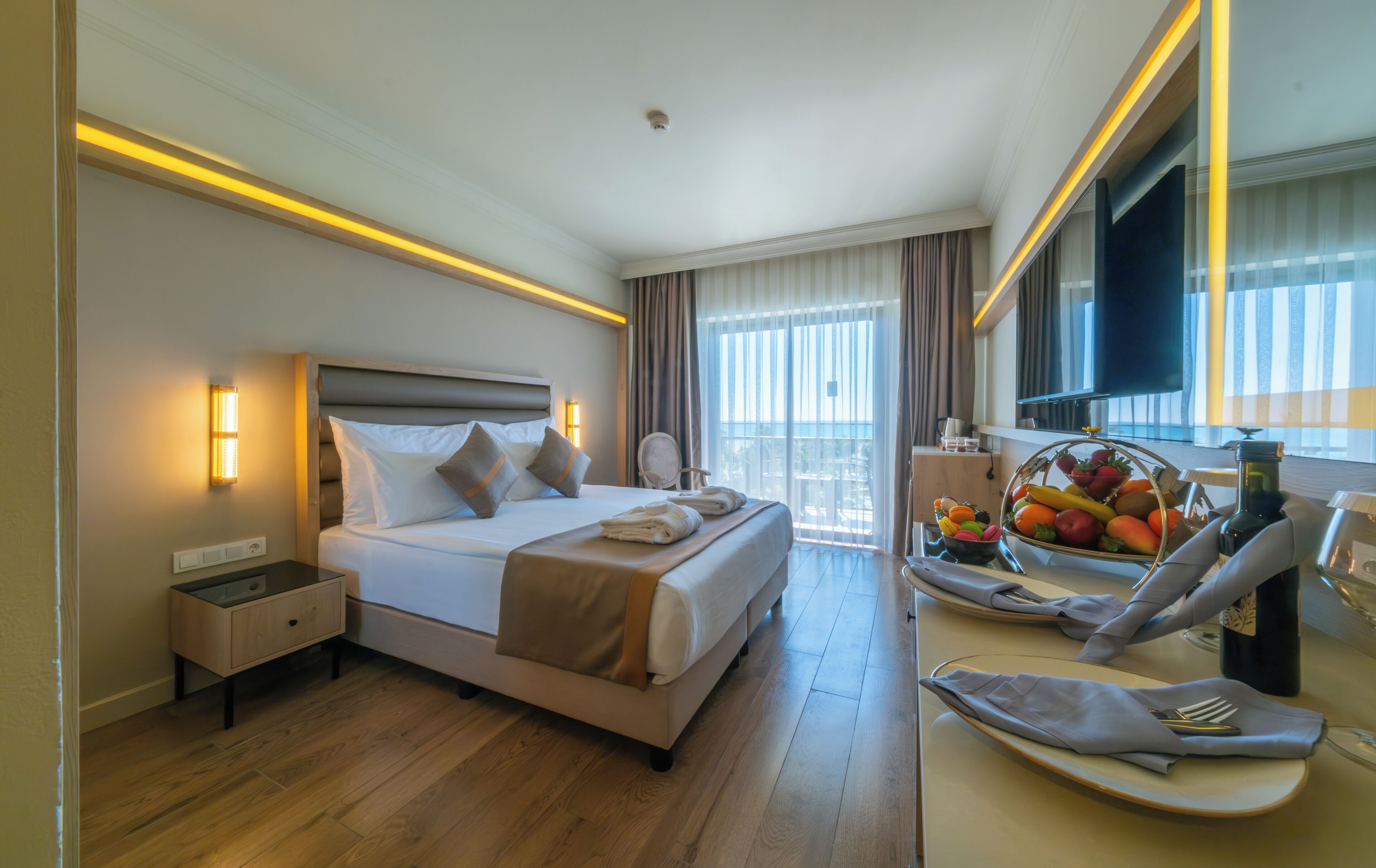 A hotel room with a large bed, bedside tables, a desk with a chair, a fruit basket, and a TV.