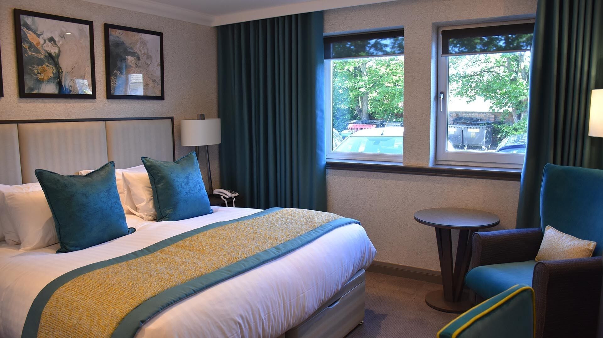 A hotel room with a large bed, a small table, and two chairs. There are three framed pictures on the wall above the bed.