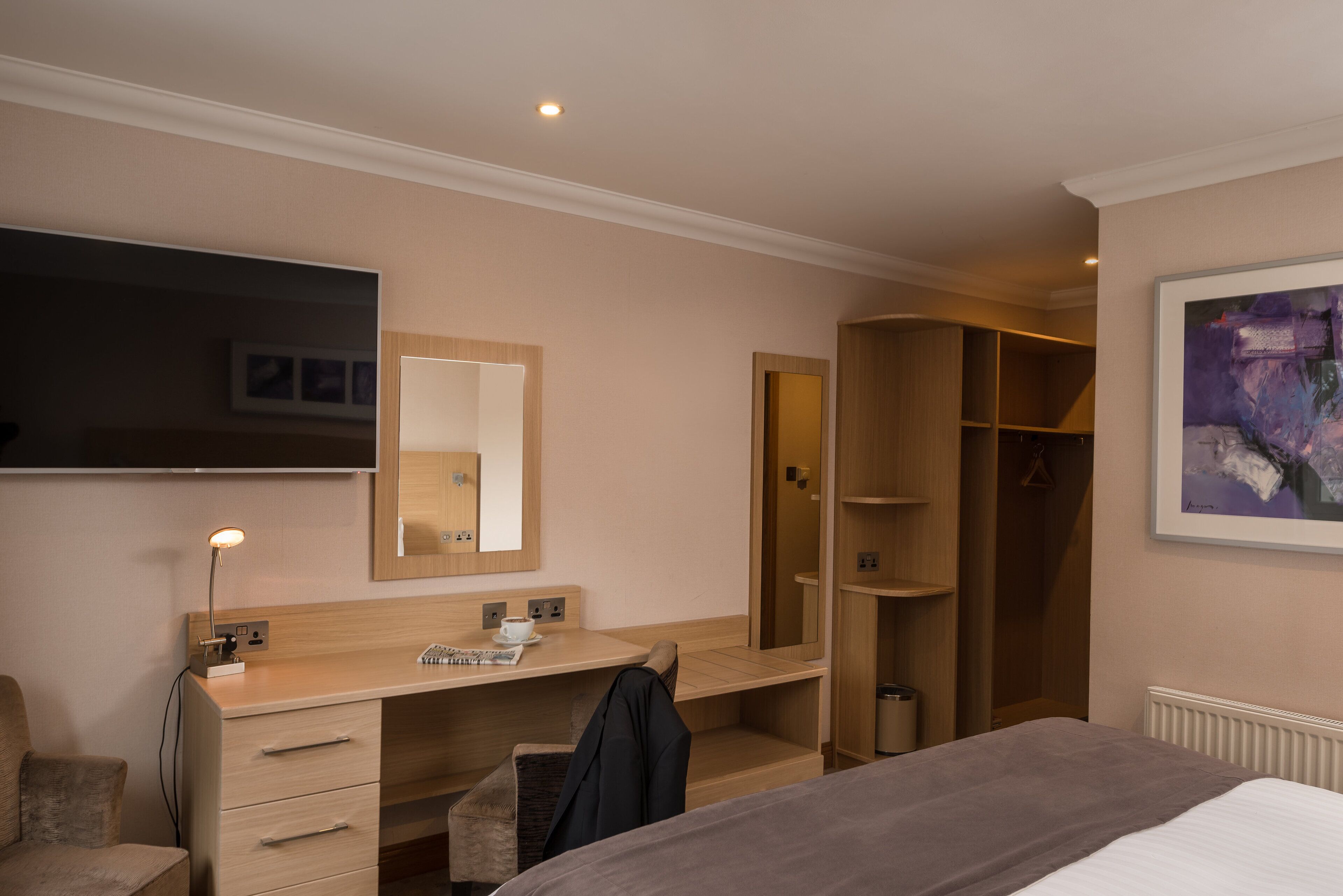 Una habitación de hotel con una cama, un escritorio con silla, un espejo, un televisor y un armario.