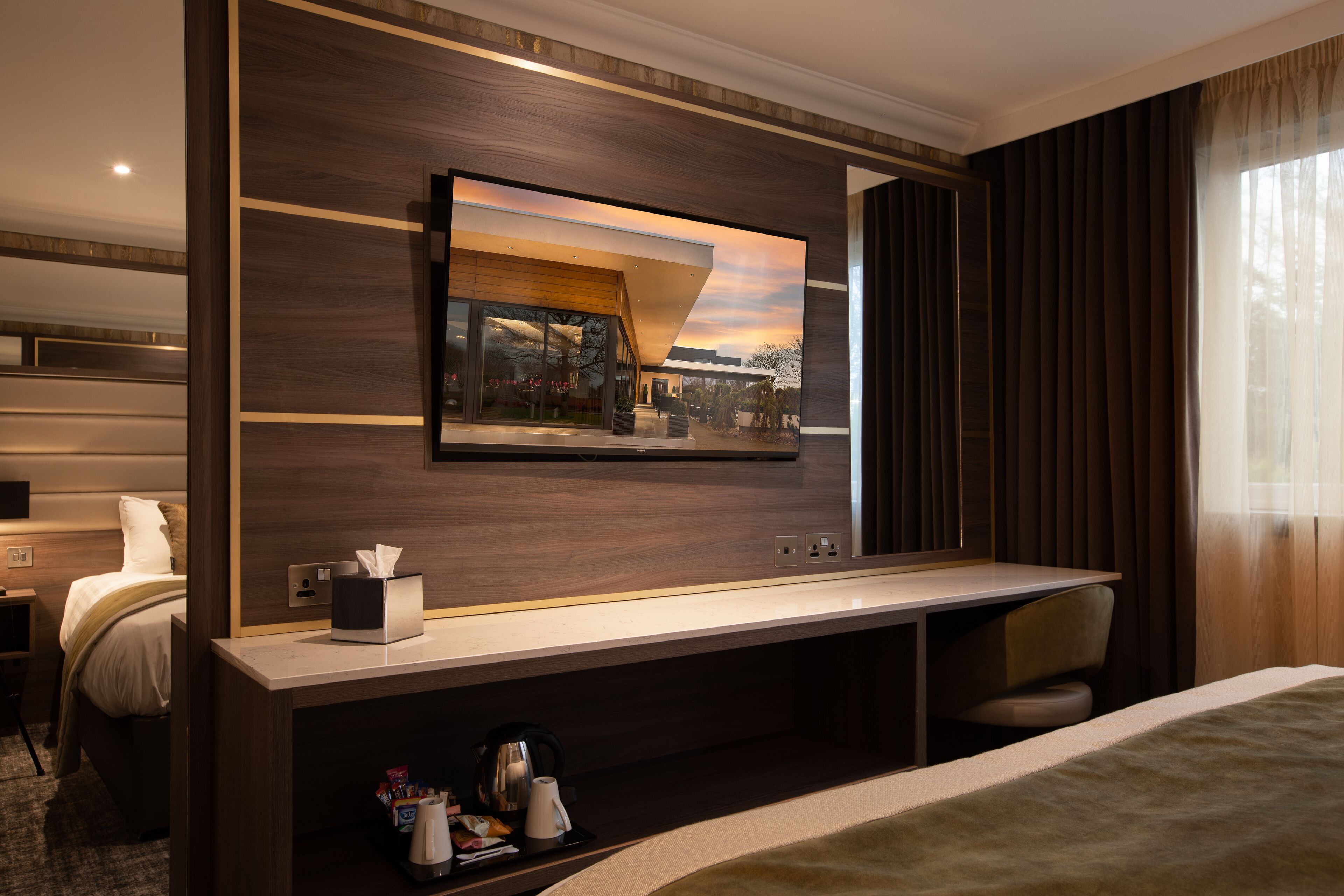 A modern hotel room with a large bed, a desk with a mirror, and a view of the outdoors.
