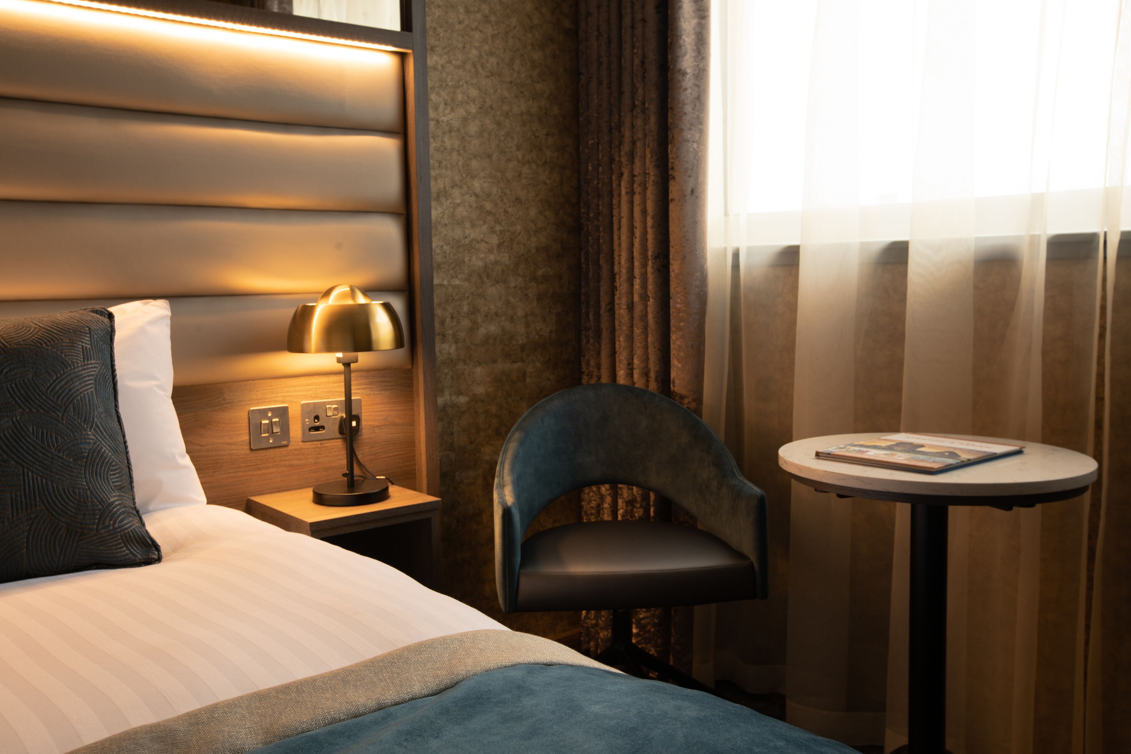 Une chambre d’hôtel avec un lit, une table de chevet avec une lampe, une chaise et une petite table sur laquelle se trouve un livre.
