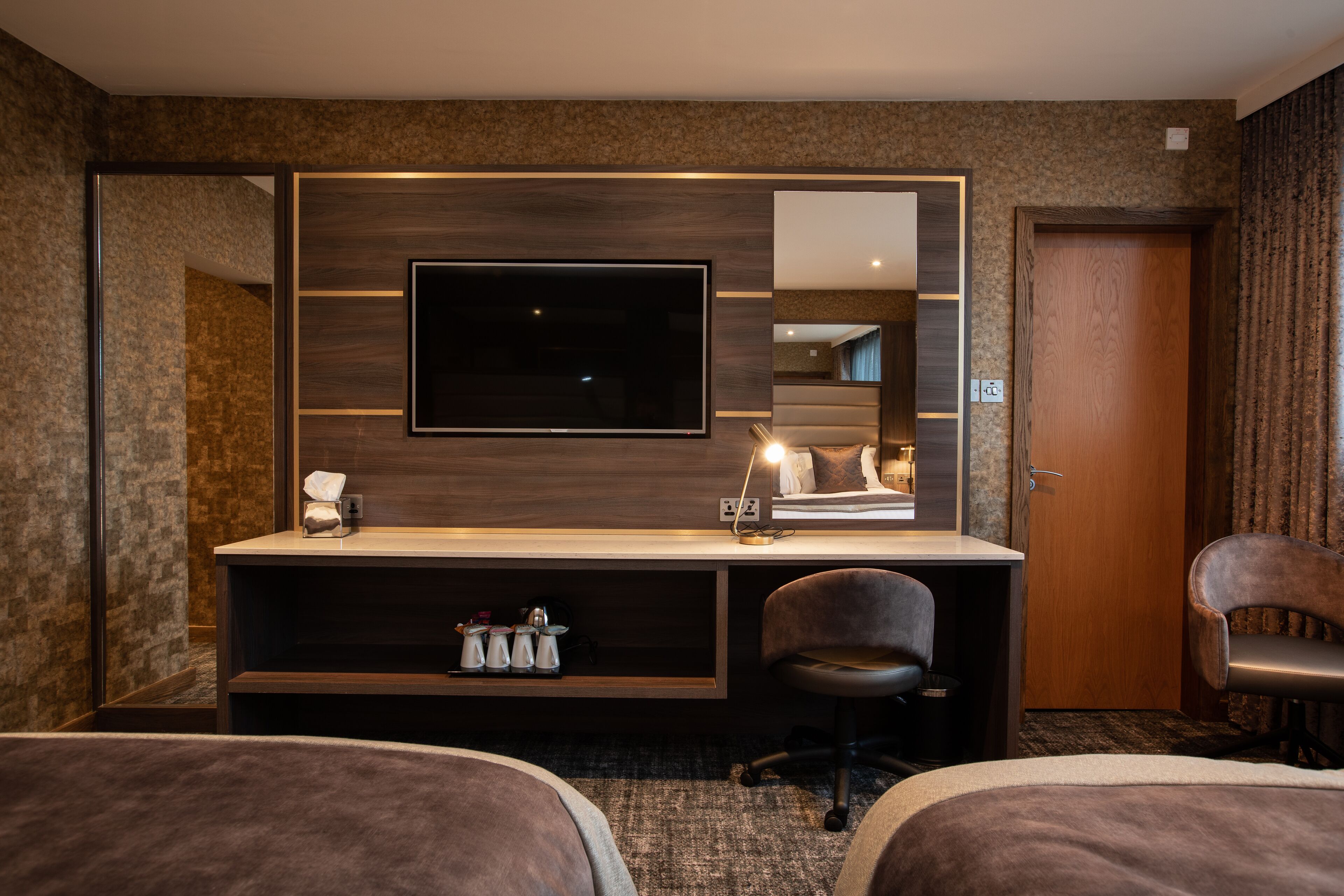 Ein Hotelzimmer mit großem Flachbildfernseher an einer Holzwand, einem Schreibtisch mit Stuhl, einem Bett und einer Tür mit Spiegel.