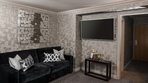 A living room with a dark velvet sofa, patterned cushions, a wall-mounted TV, and a small table with a bottle and glasses.