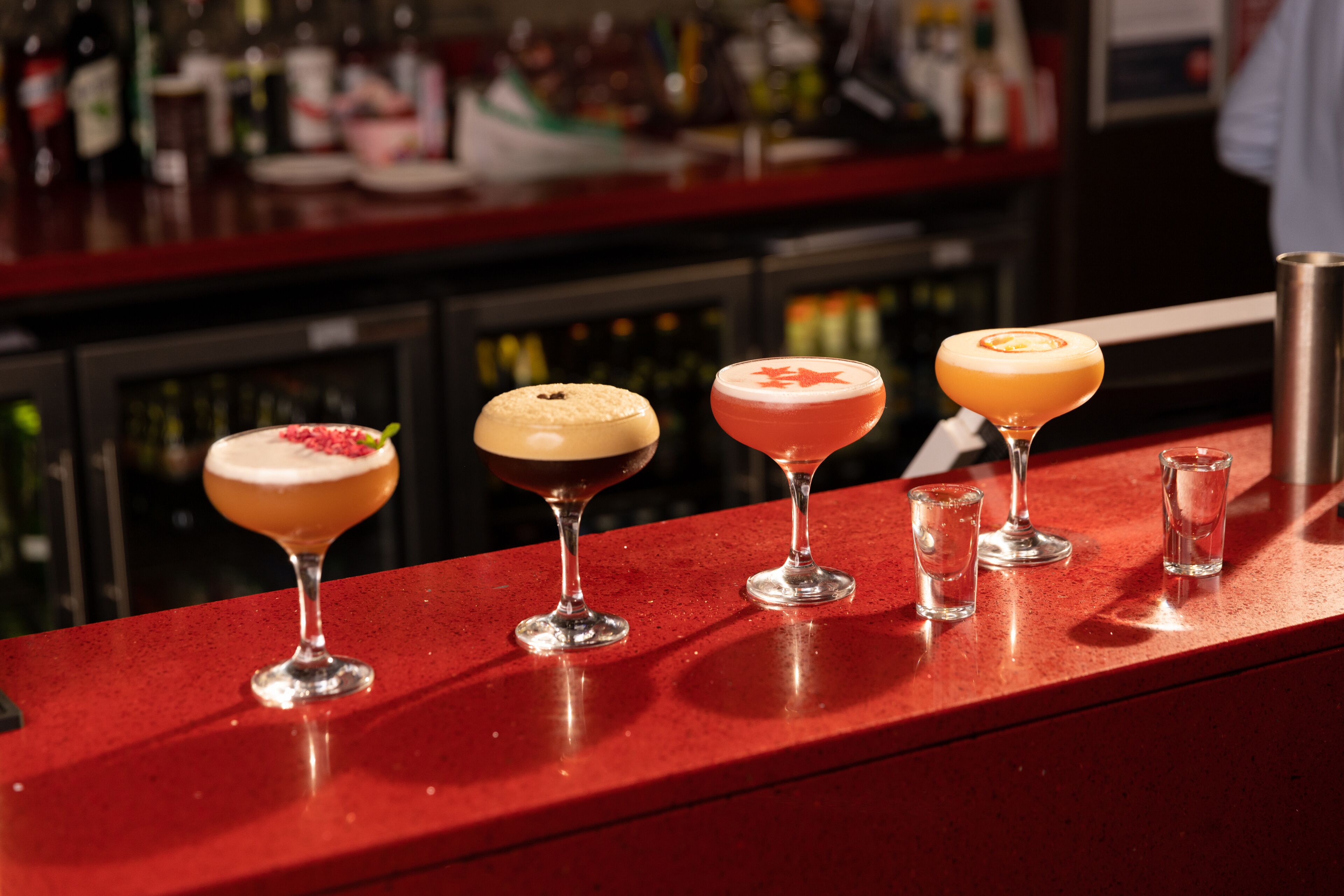 Una barra con cuatro cócteles diferentes y algunos vasos para chupitos.