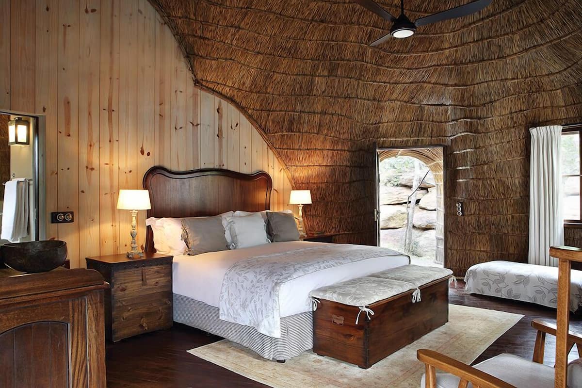 Ein Schlafzimmer mit Holzboden, einem großen Bett mit Kopfteil, Nachttischen mit Lampen, einer Holzkiste und Blick ins Freie durch eine Tür.