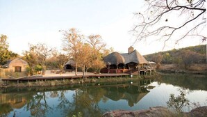 Een rustiek vakantiehuis met rieten dakbungalows aan een kalm meer, omringd door bomen en een heldere lucht.
