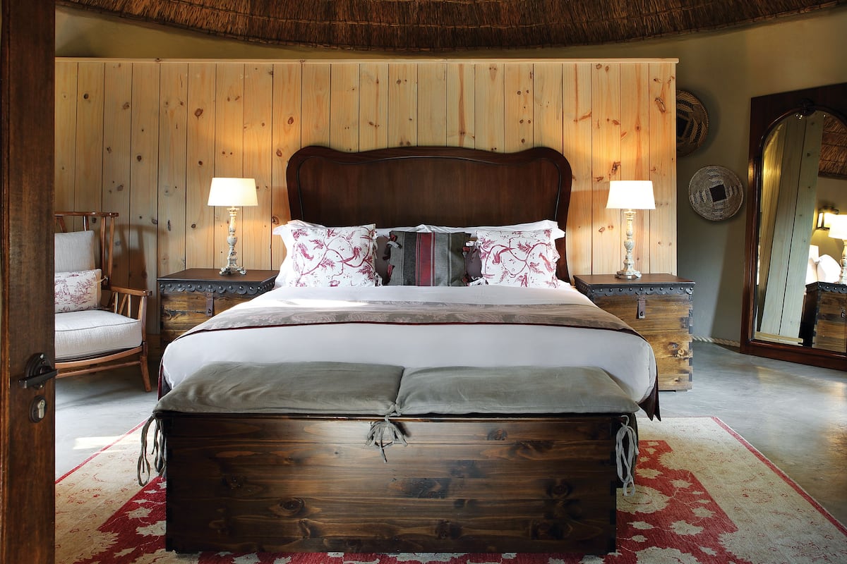 Ein hölzernes Bett mit Kopfteil, zwei Nachttische mit Lampen, ein Sessel und ein Spiegel an der Wand.
