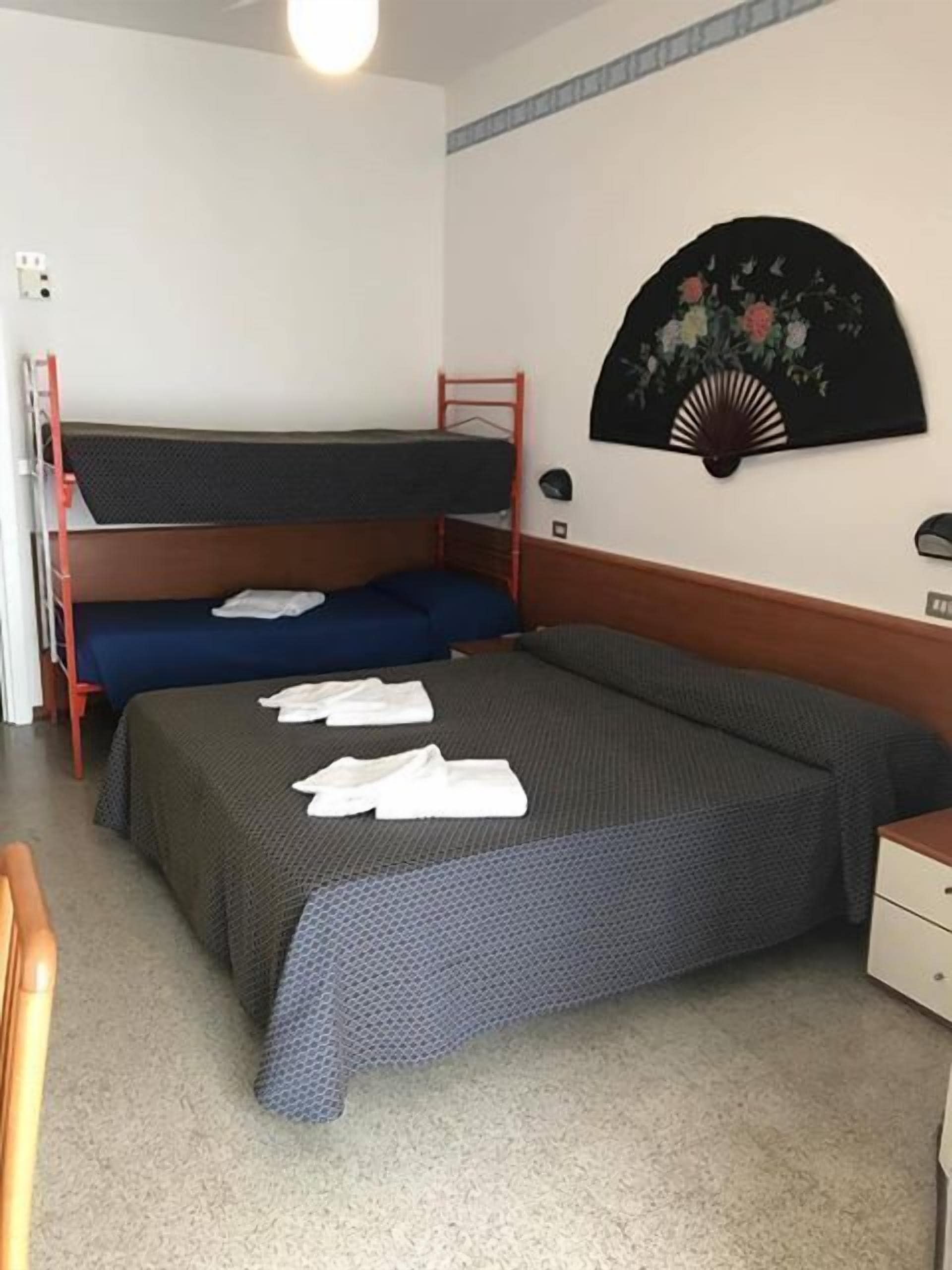 Ein Einzelbett mit grauer Bettdecke und zwei Kissen, ein Hochbett links, ein kleiner Nachttisch und ein Wandventilator mit floralem Muster.