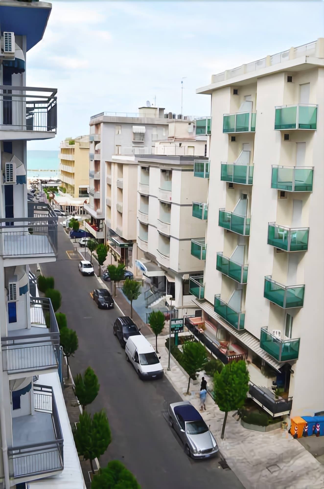A residential street with multi-story buildings, balconies, parked cars, and a pedestrian.