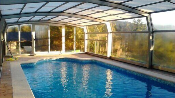 A covered swimming pool with glass walls, a clear blue pool, and a view of trees outside. - Casa Rural O'Pozo (Cerdedo-Cotobade)