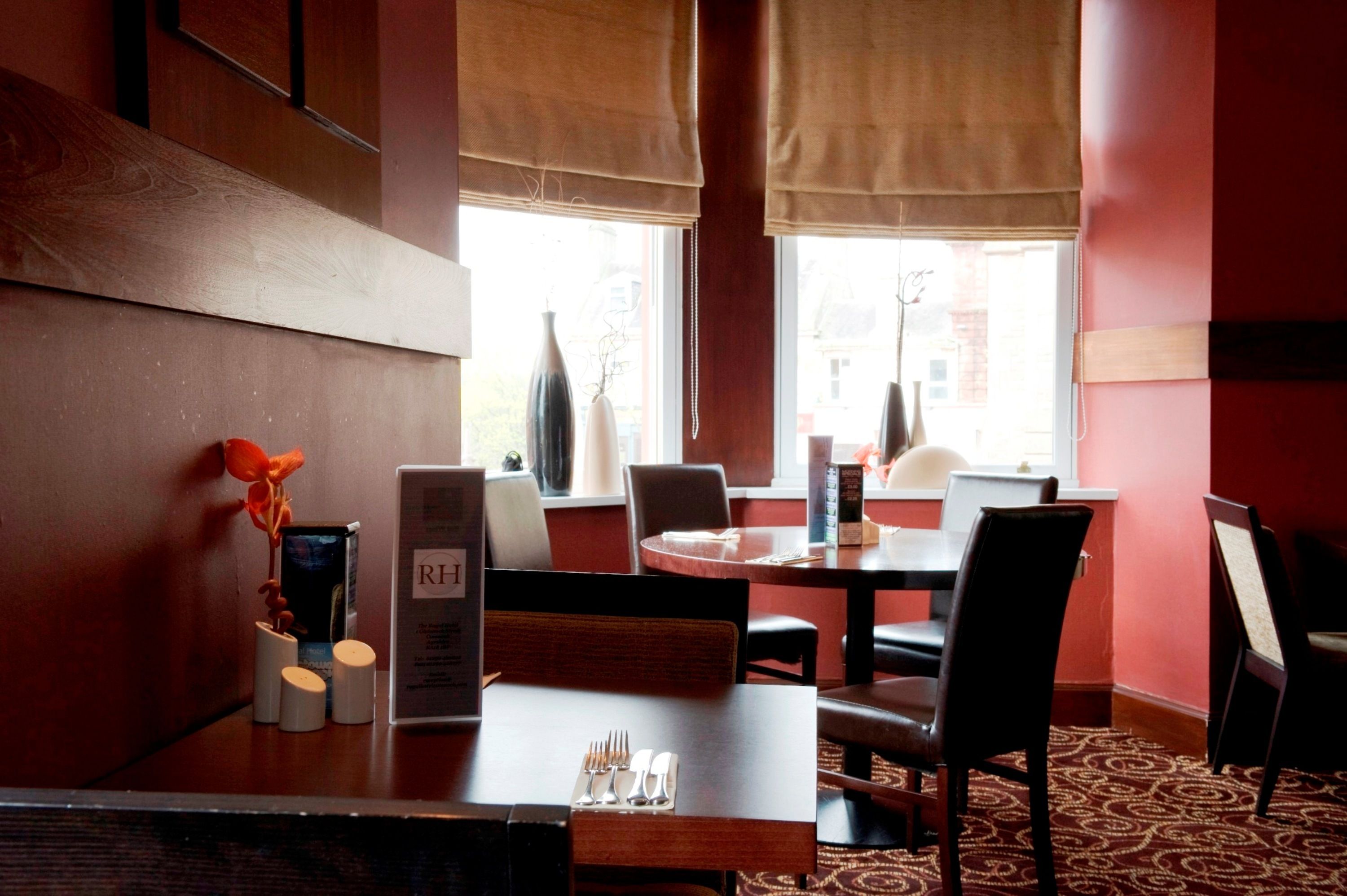 Um restaurante com interior em madeira, paredes vermelhas e grandes janelas com persianas. Mesas postas com talheres e cardápios.