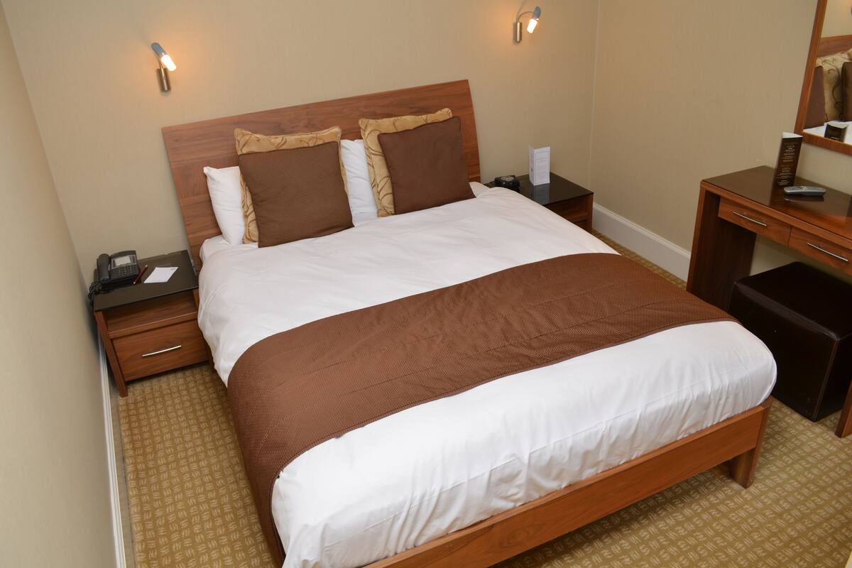 Une chambre d’hôtel avec un lit en bois, deux tables de chevet, une petite table, un bureau et un miroir.