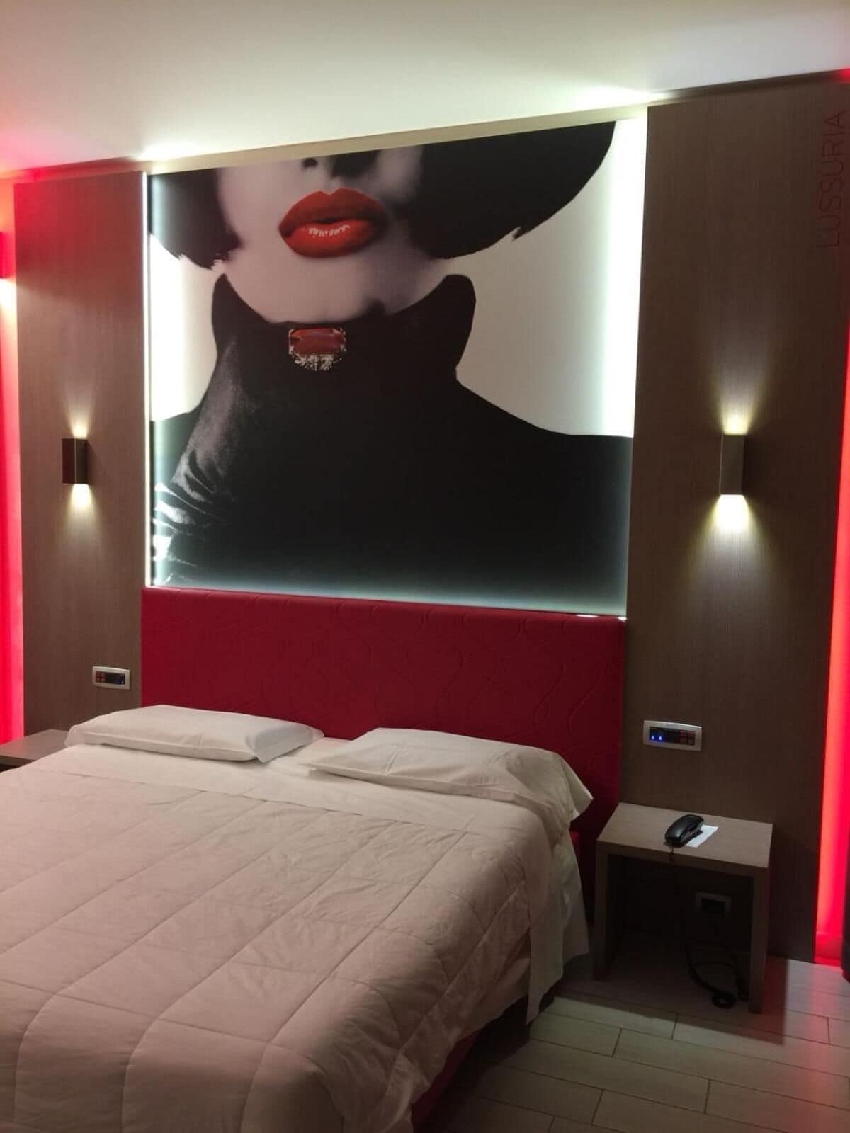 A hotel room with a large bed, a red headboard, a wall-mounted artwork, and a bedside table with a phone and a remote control.