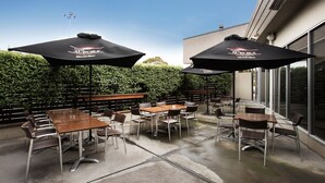 An outdoor patio with tables and chairs, two umbrellas, and a hedge in the background. - Nightcap at Gateway Hotel (Corio)