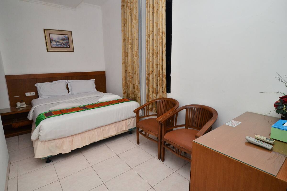 A hotel room with a bed, a wooden chair, a desk with a remote control, and a window with curtains.
