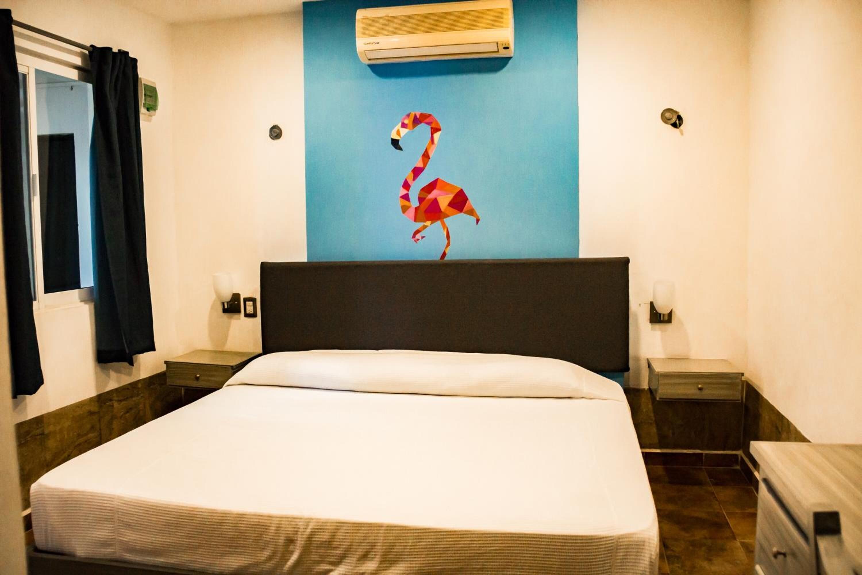 A hotel room with a bed, a nightstand, a wall-mounted air conditioner, and a wall art of a flamingo.