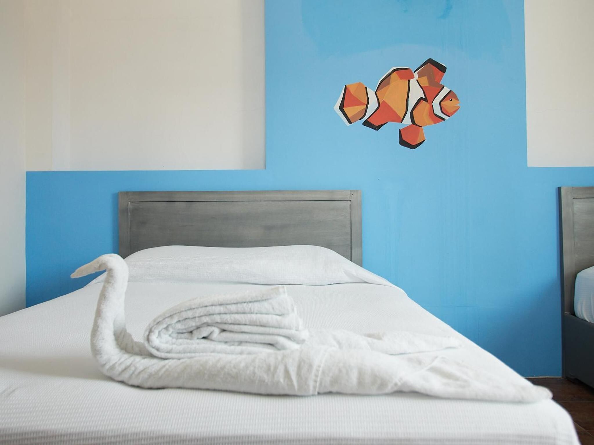 Ein ordentlich bezogenes Bett mit einem gefalteten weißen Handtuch, eine blaue Wand mit einem Fischgemälde und ein graues Kopfteil.