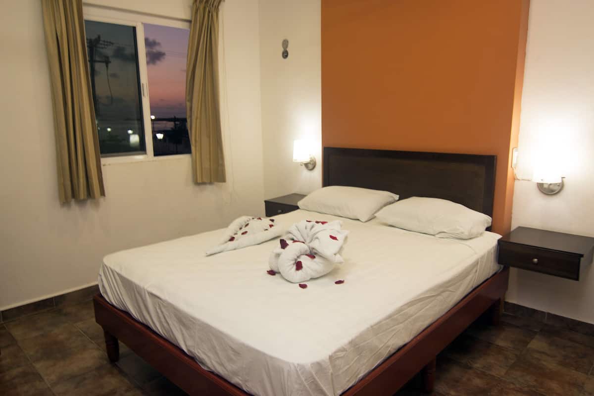 A neatly made bed with white linens and a heart-shaped towel, two bedside tables with lamps, a window with curtains, and a wall with a light switch.