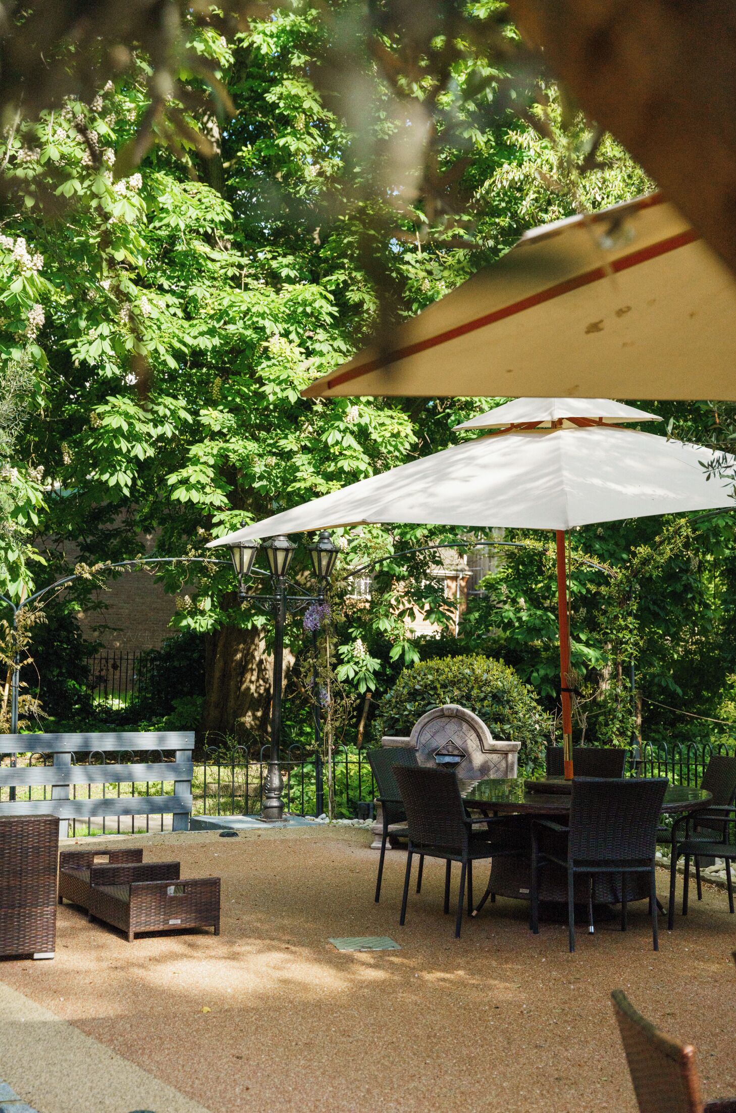 An outdoor patio with a table and chairs under a white umbrella, surrounded by greenery and a brick wall.