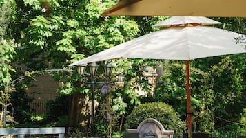 An outdoor patio with a table and chairs under a white umbrella, surrounded by greenery and a brick wall.