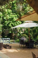 An outdoor patio with a table and chairs under a white umbrella, surrounded by greenery and a brick wall.